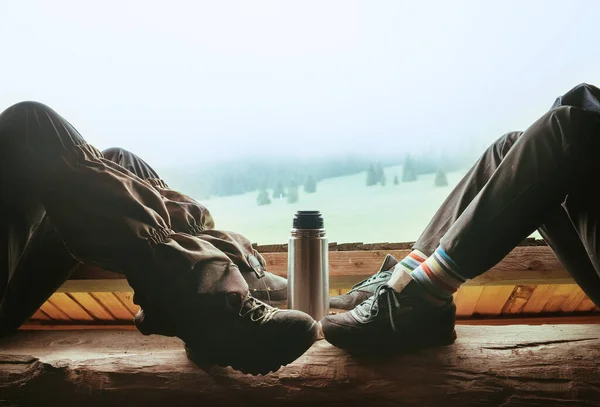 在青少年小伙子们的腿之间 他们一边喝酒 一边坐在木制的森林房子的阳台上 享受着清晨云雾密布的森林 热茶保温瓶 秋天的旅游和旅游 — 图库照片