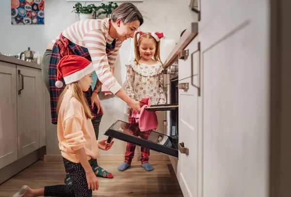 可爱的小女孩戴着红色的圣诞老人帽 妈妈做自制的甜饼干 他们把烤好的面包放在热烤箱里 圣诞饼干准备好了甜蜜的家温暖的家庭生活时刻概念 — 图库照片