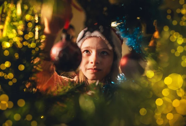 赤いサンタの帽子のかわいい女の子は おとぎ話のXマス願い事の夜にクリスマスモミの木に魔法の木の装飾ボールをぶら下げています 枝内の魅力的な子供時代と冬の休日のコンセプトイメージ — ストック写真
