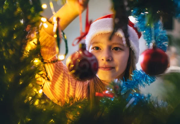 可爱的小女孩戴着红色的圣诞老人帽 挂着一个神奇的圣诞树装饰球 挂在圣诞树上 童话般的许愿之夜 迷人的童年和寒假的概念内在的分支形象 — 图库照片