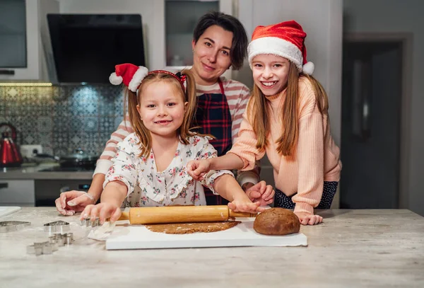 笑着带着红色圣诞礼帽的小女孩和妈妈家的波特拉特 他们在家里的厨房里用滚动式别针一起做圣诞姜饼 欢乐假期准备和童年概念 — 图库照片
