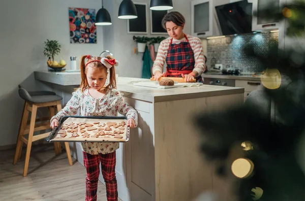 Christmas cookies preparation. Cute little girl with baking sheet full of homemade sweet ginger biscuits. She and mother preparing a celebration.  Home sweet home warm family life moments concept.