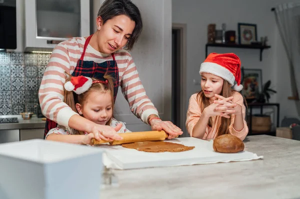 快乐地微笑着 戴着红色圣诞帽的小女孩们 妈妈在家里厨房一起揉搓圣诞姜饼面团 欢乐假期准备和童年概念 — 图库照片