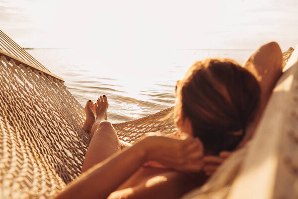 Young woman relaxing in wicker hammock on the sandy beach on Mauritius coast and enjoying sunset light over Indian ocean waves. Exotic countries vacation and mental health concept image. 