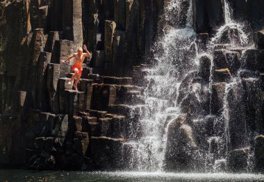 Middle-aged man jumping in waterfall lake. Falling water streams flow on black volcanic stone cascades. Rochester Falls waterfall - popular tourist spot in Savanne district in Mauritius.