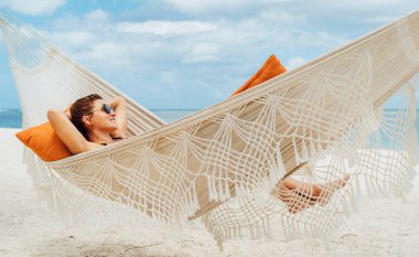 Young woman relaxing in wicker hammock on the sandy beach on Mauritius coast and enjoying wide ocean view waves. Exotic countries vacation and mental health concept image.