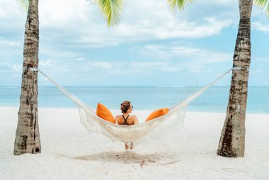 Young woman relaxing in wicker hammock on the sandy beach on Mauritius coast and enjoying wide ocean view waves. Exotic countries vacation and mental health concept image.
