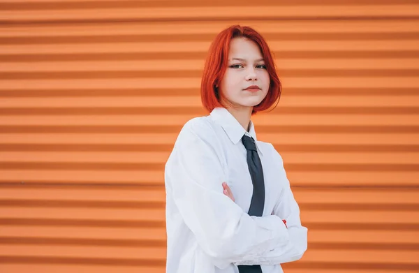 Bright portrait of teenage girl with painted red dyed hair in white school shirt and necktie standing near orange wall background with hands crossed body posture.