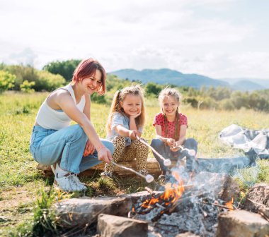 Three sisters cheerfully smiling while they roasting a marshmallows candies on the sticks over the campfire flame. Happy family or outdoor picnic activities concept image.	