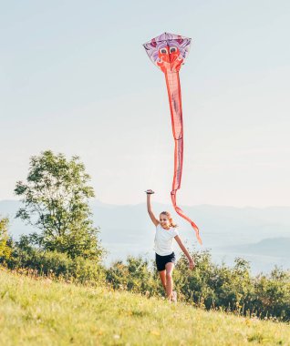 Little girl with flying a colorful kite on the green grass meadow in the mountain fields. Happy childhood moments or outdoor time spending concept image. 