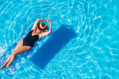 Siyah bikinili seksi bir kadın, hasır şapkalı ve güneş gözlüklü şişme yatakta yüzen mavi yüzme havuzu dalgaları üzerinde. Lüks tatil beldelerinde yaz tatili konsepti..