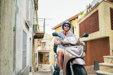 Kasklı ve güneş gözlüklü neşeli bir adam dar Sicilya eski sokaklarında moto scooter kullanıyor. Mutlu İtalyan tatili ve ulaşım konsepti.