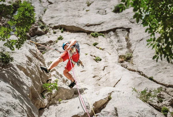 活跃的登山者中年男子戴着防护头盔 用绳索和系带装置从悬崖岩壁上爬下 积极的极限运动时间概念 — 图库照片