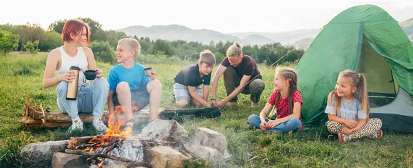笑顔の子供たちのグループは 煙の多いキャンプファイヤーの近くで陽気な会話をしています 彼らは魔法瓶からお茶を飲む 2人の兄弟は緑のテントを設定します 幸せな家族の屋外ピクニックキャンプ活動のコンセプト — ストック写真
