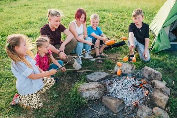 6人の子供たちグループ男の子と女の子は 緑のテントの近くのキャンプファイヤーの炎の上に棒で陽気に笑ってローストソーセージ アウトドアアクティブ時間自然コンセプトで過ごすか キャンプ — ストック写真