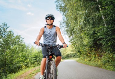 Portrait of happy man dressed in cycling clothes, helmet and sunglasses riding a bicycle on the out-of-town bicycle path. Active sporty people concept image.	