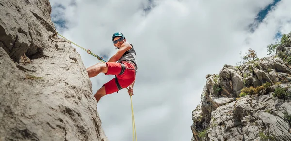 活跃的登山者中年男子戴着保护头盔 用绳索 腰带装置和攀爬带 从悬崖岩墙上垂下时看着相机 积极的极限运动时间概念 — 图库照片
