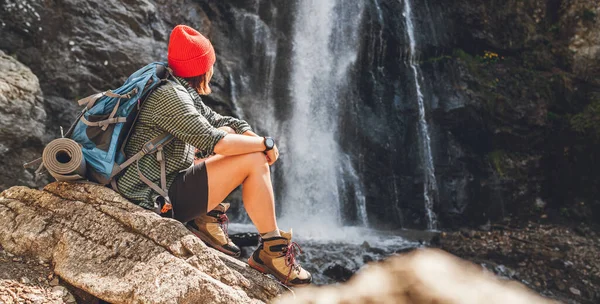 一个戴着红帽子背着背包的女人 穿着活跃的步行服 坐在山河瀑布边 享受着大自然的力量 远足和自然概念形象 — 图库照片