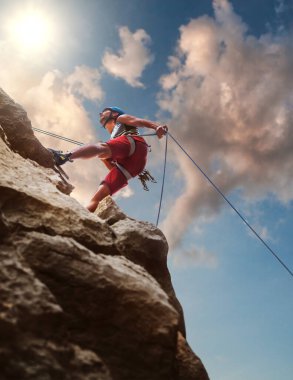 Koruyucu miğferli kaslı adam kayalık duvardan halatla iniyor ve akşamüstü gökyüzü arka planında tırmanma kemeri kullanıyor. Aktif ekstrem spor zaman harcama kavramı.