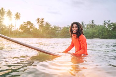 Uzun sörf tahtasının üzerinde yüzen siyah uzun saçlı genç adam palmiye koruluğuyla sörf yapmaya hazır bir dalga bekliyor. Ekstrem su sporları ya da egzotik ülkelere seyahat. Sri Lanka