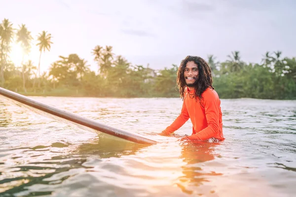 stock image Black long-haired teen man floating on long surfboard, waiting for a wave ready for surfing with palm grove litted sunset rays. Extreme water sports or traveling to exotic countries concept. Sri Lanka