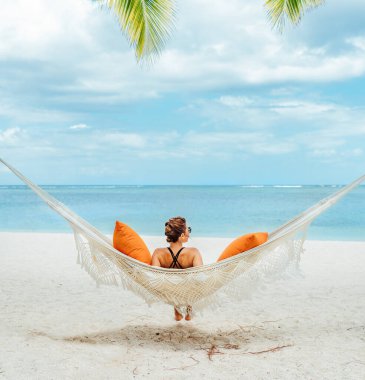 Young woman relaxing in wicker hammock on the sandy beach on Mauritius coast and enjoying wide ocean view waves. Exotic countries vacation and mental health concept image.