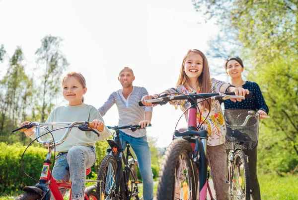 夏天外出骑自行车时 带着两个女儿微笑的父母 他们在夏季城市公园里聚精会神 快乐生儿育女或积极的体育生活概念形象 — 图库照片