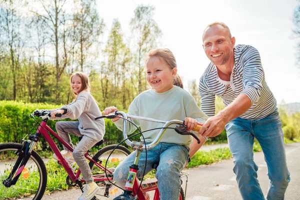 在户外散步的时候 带着两个女儿微笑的父亲 他教小女孩骑自行车 他们在夏季城市公园里聚精会神 快乐童年的概念形象 — 图库照片