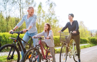 Yazın bisiklet sürerken kızıyla birlikte gülümseyen anne ve baba. Onlar yaz şehir parkında birliktelikten hoşlanırlar. Mutlu ebeveynlik ve çocukluk veya aktif spor hayatı konsepti imajı.