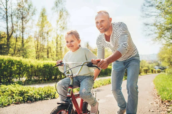 笑顔の父は自転車に乗るように娘に教える 彼らは夏の都市公園で一緒に楽しむ 幸せな子供時代のコンセプトイメージ — ストック写真