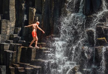 Caucasian man in swimsuit balancing near the falling water streams flowing on black volcanic stone cascades. Rochester Falls waterfall - popular tourist spot in Savanne district in Mauritius.