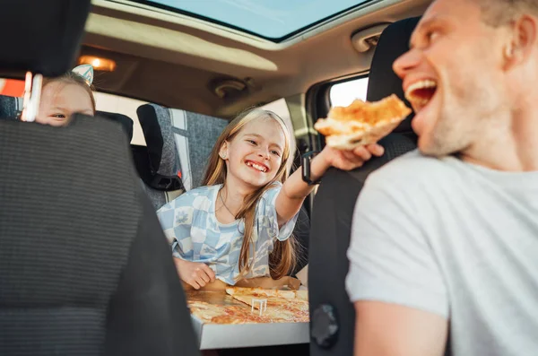 チャイルドシートに座りながらお父さんの口の中にピザを入れてポジティブな笑顔の女の子の肖像 幸せな子供時代 ファーストフードやオートジョーニーランチ休憩のコンセプトイメージ — ストック写真