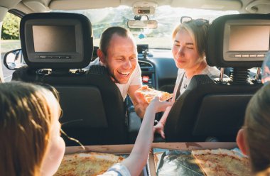 Young couple with daughters eating just cooked Italian pizza sitting in modern car with transparent roof. Happy family moments, childhood, fast food eating or auto journey lunch break concept image.	