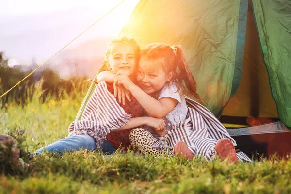 2人の小さな抱擁の女の子の緑の芝生のキャンプテントの入り口の横に座って 陽気に笑みを浮かべて 不注意な子供時代 家族の価値観と屋外活動のコンセプト写真 — ストック写真