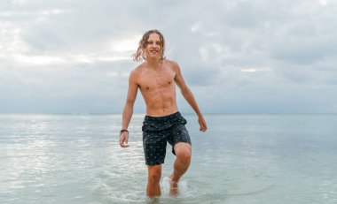 Smiling muscular longhaired teenage boy walking out from Indian ocean waves after evening swimming. Traveling and an exotic vacations concept image.