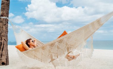 Young woman relaxing in wicker hammock on the sandy beach on Mauritius coast and enjoying wide ocean view waves. Exotic countries vacation and mental health concept image.