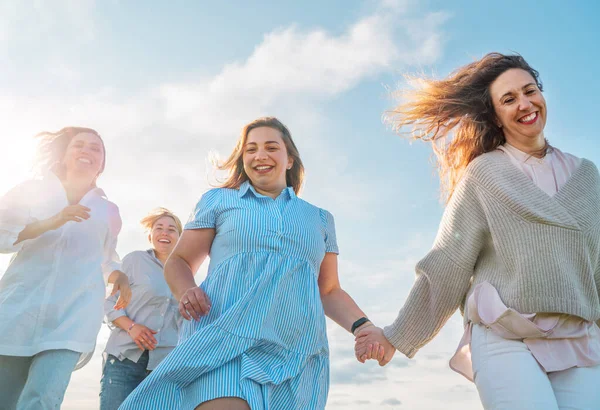 四个笑容满面的女人手牵着手在草地上奔跑的画像 用云彩在蓝天上拍摄的低角度照片 女人的友谊 关系和幸福的概念形象 — 图库照片