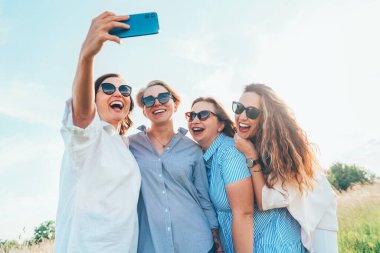 Güneş gözlüklü, gülümseyen dört kadının portresi. Birbirlerine sarılıyorlar ve doğa yürüyüşü sırasında modern akıllı telefon kullanarak selfie çekiyorlar. Kadınların dostluğu, ilişkileri ve mutluluk konsepti imajı