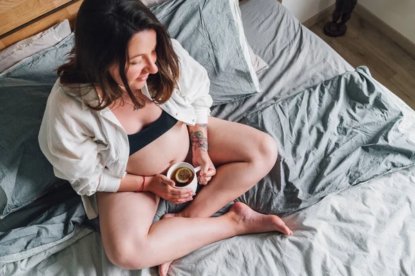 若い妊婦のクロスに座って ベッドルームでお茶のカップとベッドの上に早朝の時間に足 女性の健康 幸せな妊娠と穏やかな精神的な気分の概念イメージ — ストック写真