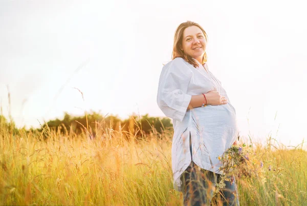 真剣に笑顔若い妊婦の光の夏の服を夕方の日没の時間に野の花の花束で高い緑の芝生の牧草地を歩いて身を包んだ 自然界の概念イメージにおける人間 — ストック写真