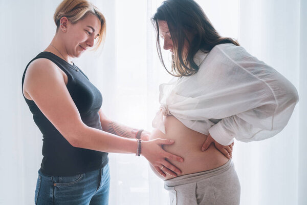 Young woman tender touching partner's female pregnant belly. Same-sex marriage couple next to living room window. Woman's health, happy pregnancy doula supporting and calm mental mood concept image.
