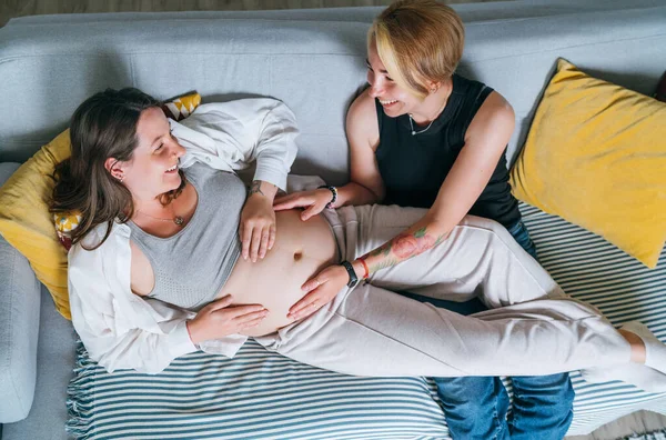 年轻女子轻柔触摸伴侣的女性怀孕腹部 同性伴侣在家里客厅的沙发上 女人的健康 快乐的怀孕 支持和镇定的心理观念形象 — 图库照片