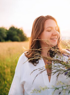 İçtenlikle gülümseyen, yaz kıyafetleri içinde genç hamile bir kadın akşam saatlerinde kır çiçekleri buketi ile yeşil çayır boyunca yürüyor. Doğa konsepti imajında insan.