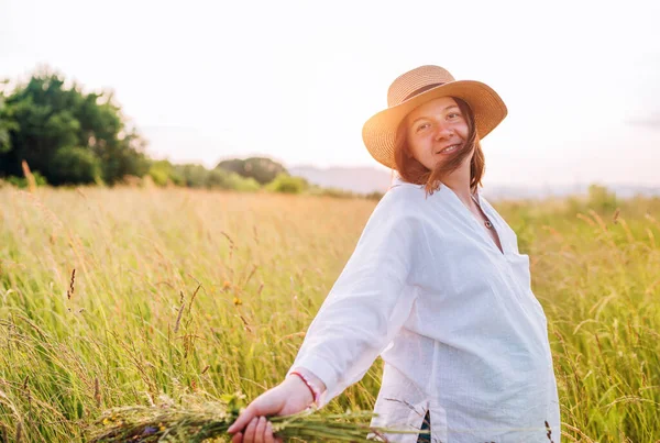 薄手の夏服に身を包んだ若い妊婦を笑顔にし 夕方の夕日の時間に野の花の花束で高い緑の草の牧草地を歩くわら帽子 自然界の概念イメージにおける人間 — ストック写真
