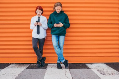 İki Kafkas ergen erkek ve bir kız, turuncu duvar arka planına yaslanırken akıllı telefonlarına göz atıyorlar. Dikkatsiz gençler. zaman ve modern teknoloji konsept görüntüsü. 