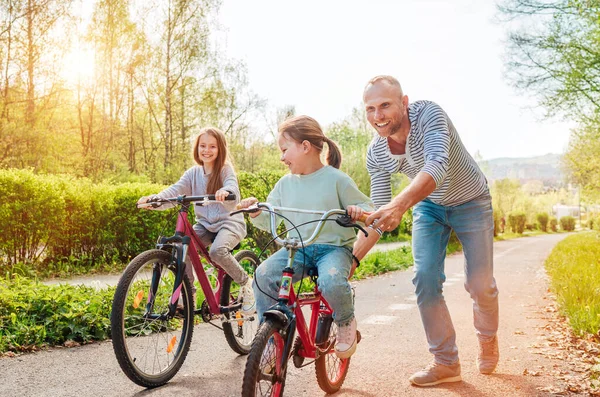 屋外の散歩中に2人の娘と笑顔の父 彼は若い女の子に自転車に乗るように教えている 彼らは夏の都市公園で一緒に楽しむ 幸せな子供時代のコンセプトイメージ — ストック写真