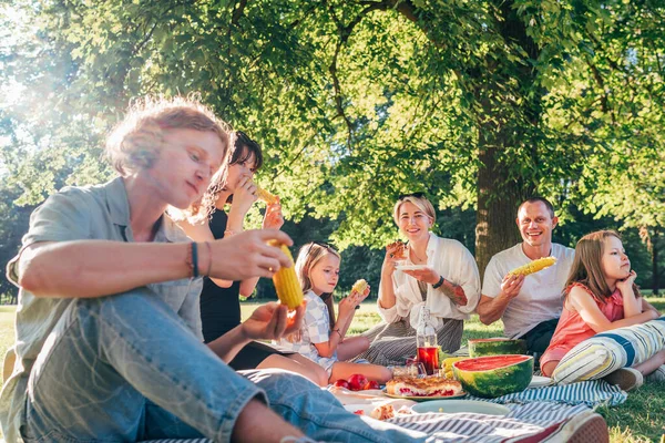 周末阳光明媚的一天 一家人坐在城市公园里的野餐毯上 他们笑着 笑着吃着煮熟的玉米和西瓜 家庭价值观和户外活动概念 — 图库照片