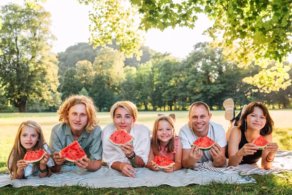 大きな家族は 週末の日曜日の晴れた日にリンデンの木の下の都市公園のピクニック毛布の上に横たわって カメラで笑って スイカを食べています 家族の価値観と野外活動の概念 — ストック写真