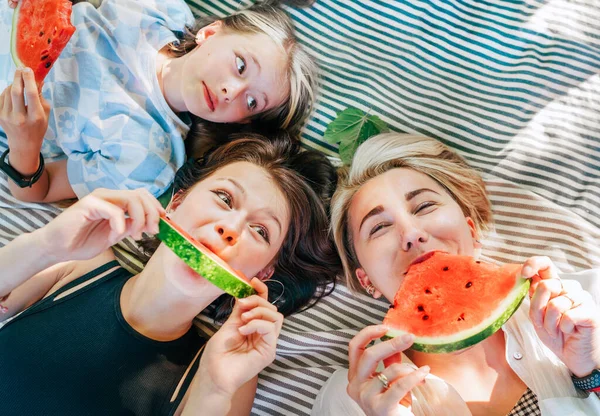周末阳光明媚的日子里 妈妈和女儿们躺在城市公园的野餐毯上 笑着相机 吃着多汁的西瓜 家庭价值观 水果维生素 户外时间观念 — 图库照片