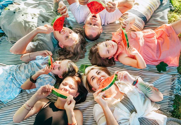 周末阳光明媚的日子 与父母团聚的女儿们躺在野餐毛毯上 笑着吃着红色多汁的西瓜片 家庭价值观 水果维生素 户外时间观念 — 图库照片